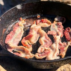 Fried bacon in a cast iron pan cooking over fire