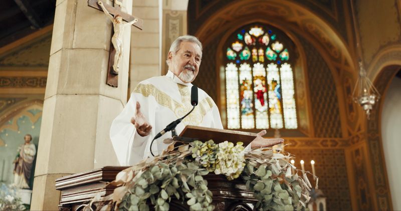 priest wearing white vestments preaching from the pulpit in a church; there is a stained glass window behind him and a crucifix, and the pulpit is decorated with flowers